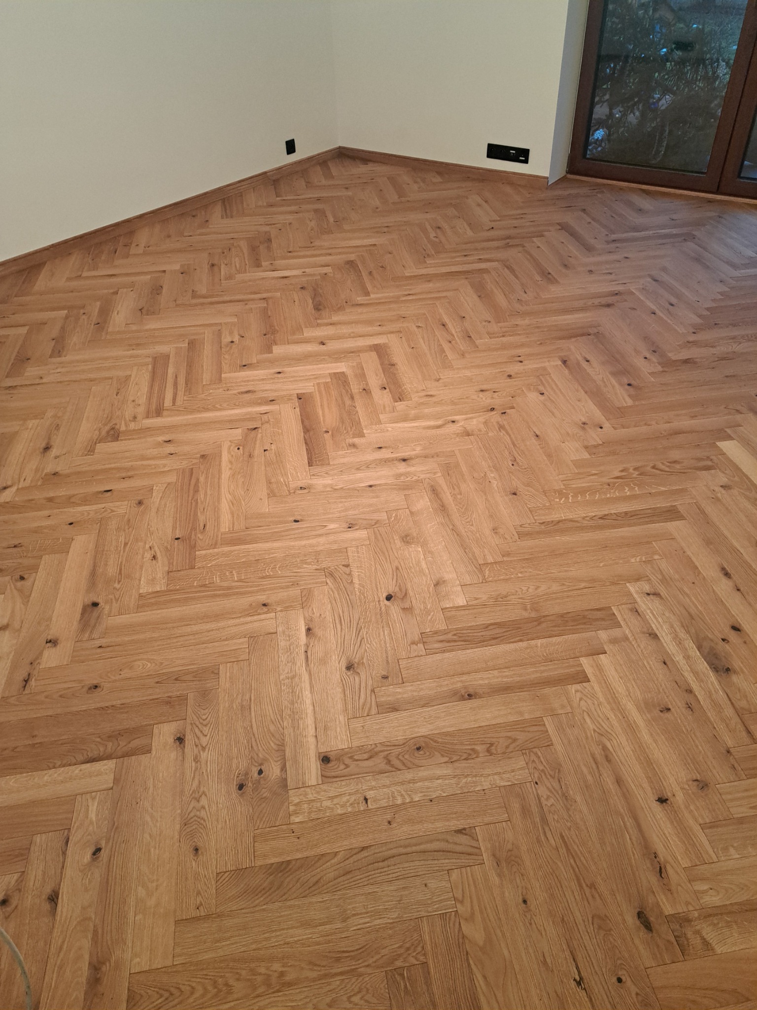 Ukończona podłoga z parkietu ułożonego we wzór jodełki francuskiej, z widocznymi naturalnymi słojami drewna i małymi sękami, przy ścianie gniazdka elektryczne.