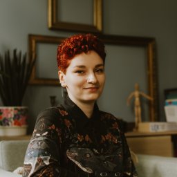 Centrum Libertus - Psychologia - Psychoterapia - Psychodietetyka - Dietetyka - Portret uśmiechniętej kobiety z krótkimi rudymi włosami, siedzącej na szarym fotelu w pokoju z ramami na ścianie i drewnianą figurką na komodzie.