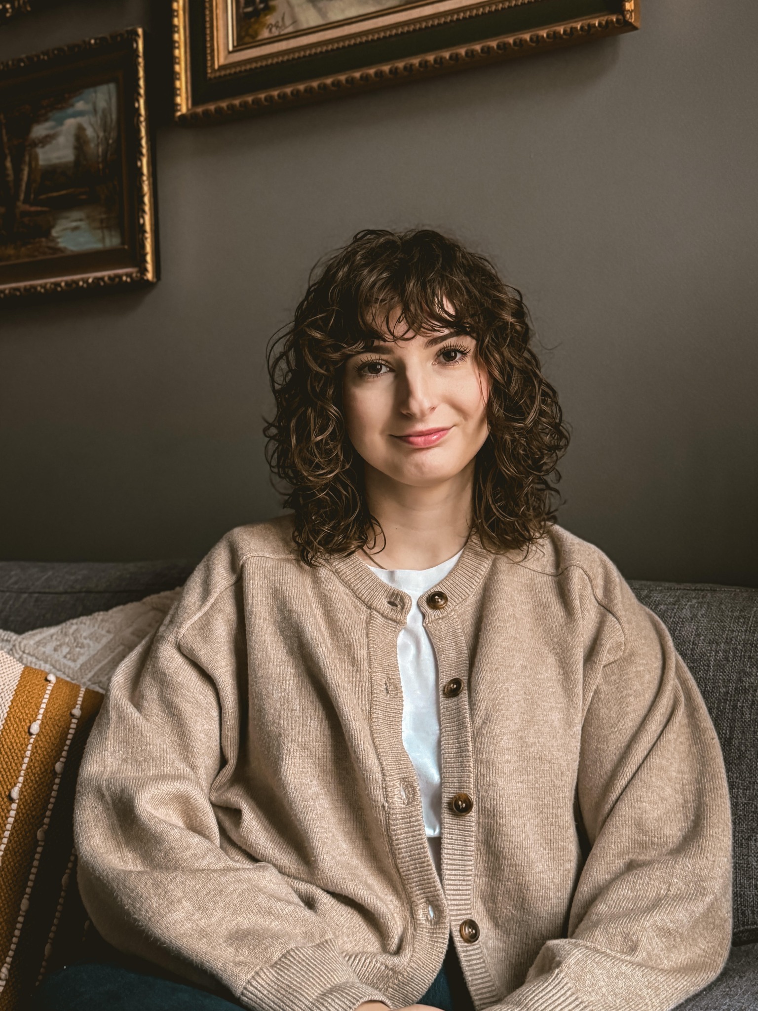 Portret uśmiechniętej kobiety o kręconych włosach w beżowym kardiganie, siedzącej na szarej sofie z dekoracyjnymi poduszkami w tle, na ścianie wiszą dwa obrazy w złotych ramach.
