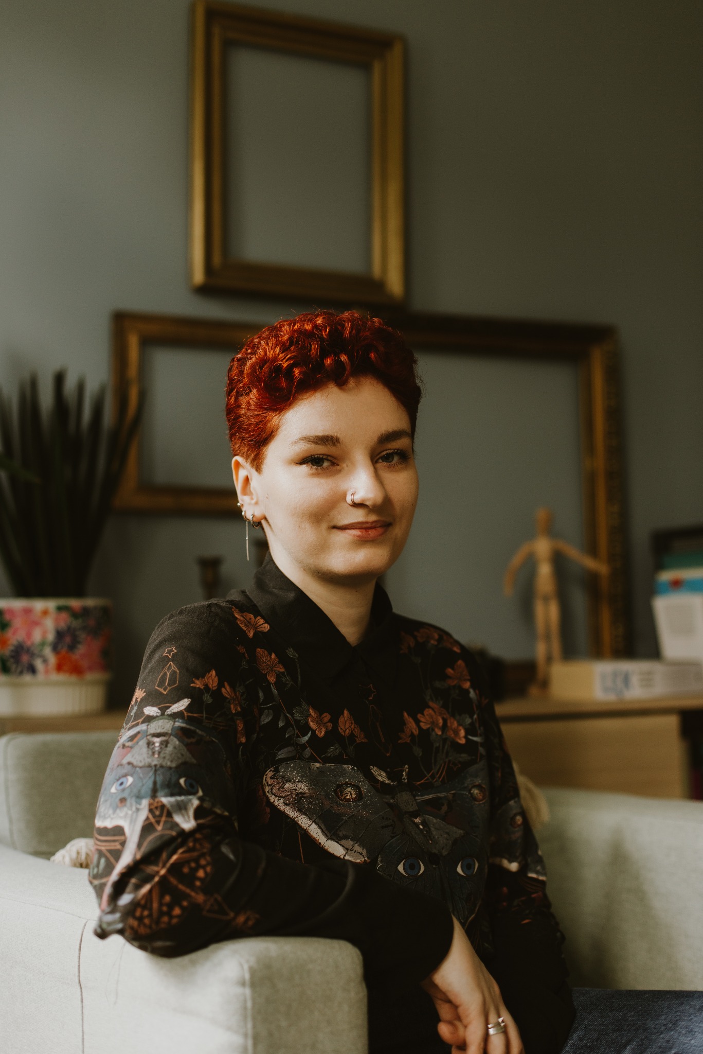 Portret uśmiechniętej kobiety z krótkimi rudymi włosami, siedzącej na szarym fotelu w pokoju z ramami na ścianie i drewnianą figurką na komodzie.