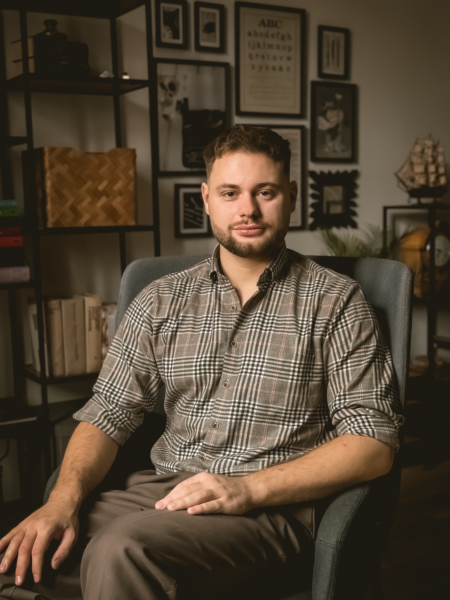Mężczyzna w kraciastej koszuli siedzi w fotelu na tle regału z książkami i dekoracjami ściennymi.