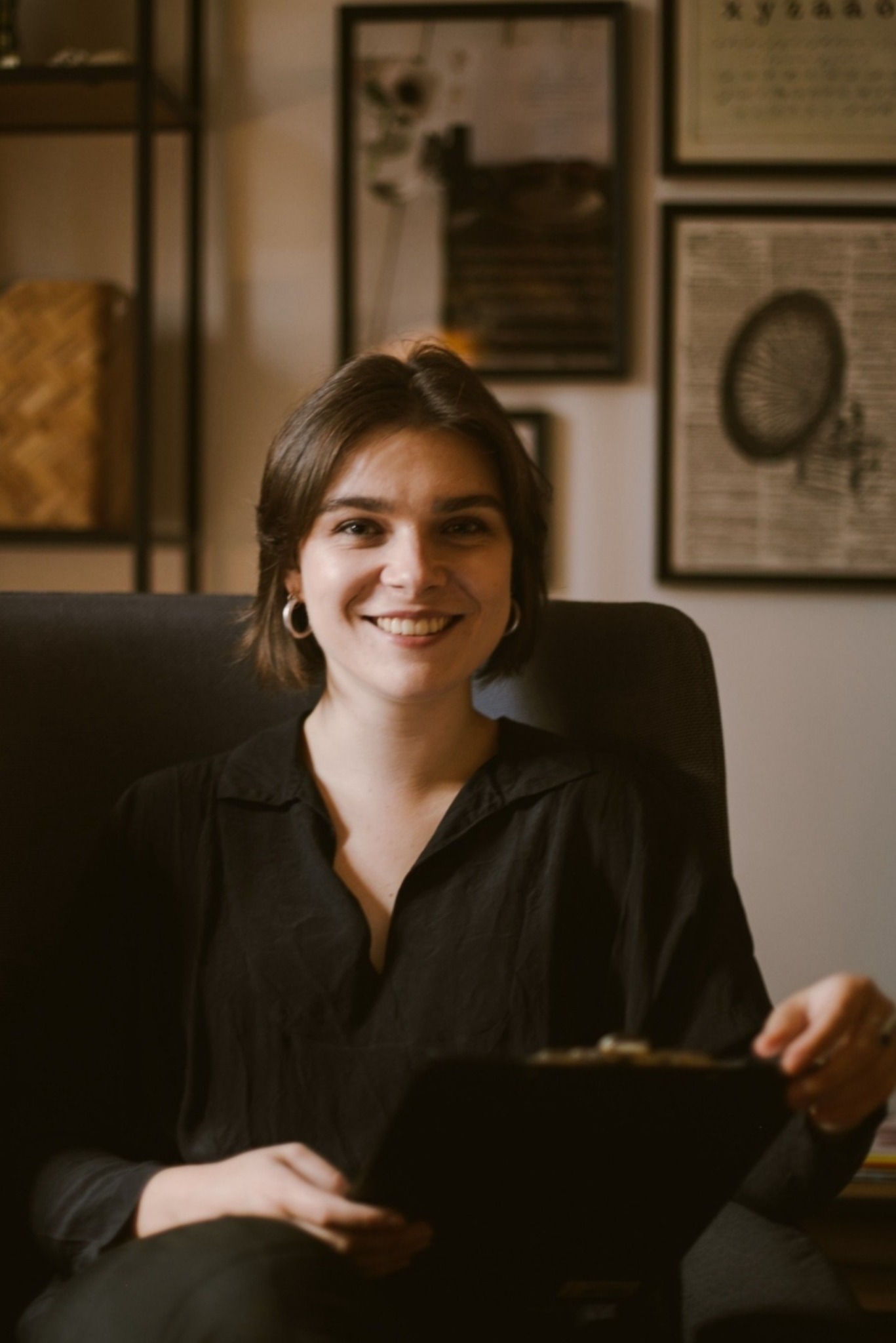 Uśmiechnięta kobieta z krótkimi brązowymi włosami siedzi w fotelu, trzymając czarną podkładkę z dokumentami; za nią widoczne półki i grafiki w ramkach.