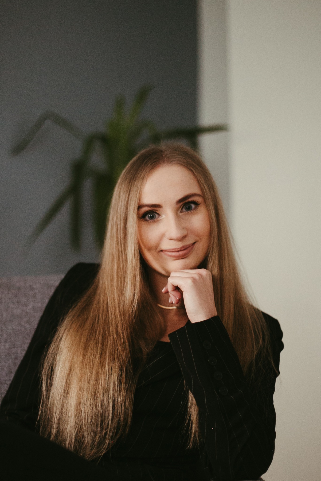 Portret kobiety z długimi blond włosami, opierającej brodę na dłoni, ubrana w czarną marynarkę w prążki, siedzącej na szarym fotelu na tle zielonej rośliny i jasnej ściany.