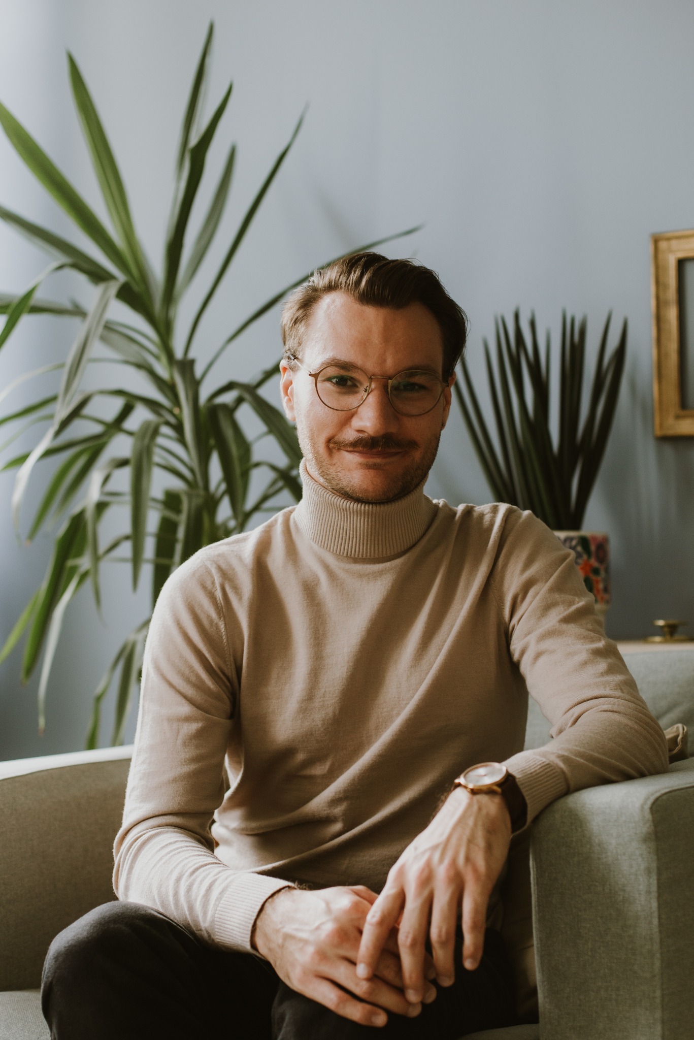 Portret mężczyzny w beżowym golfie i okularach, siedzącego na szarej sofie z rośliną w tle, prezentujący się jako psycholog lub psychoterapeuta.