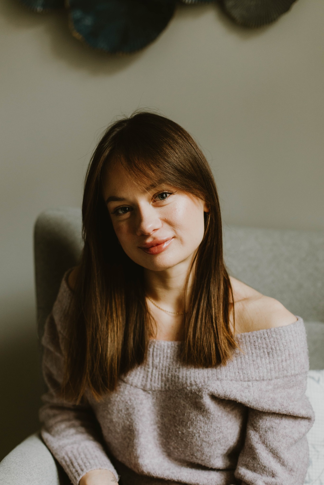 Portret uśmiechniętej kobiety z brązowymi włosami w swetrze z odkrytymi ramionami, siedzącej na szarym fotelu na tle ściany z dekoracją.