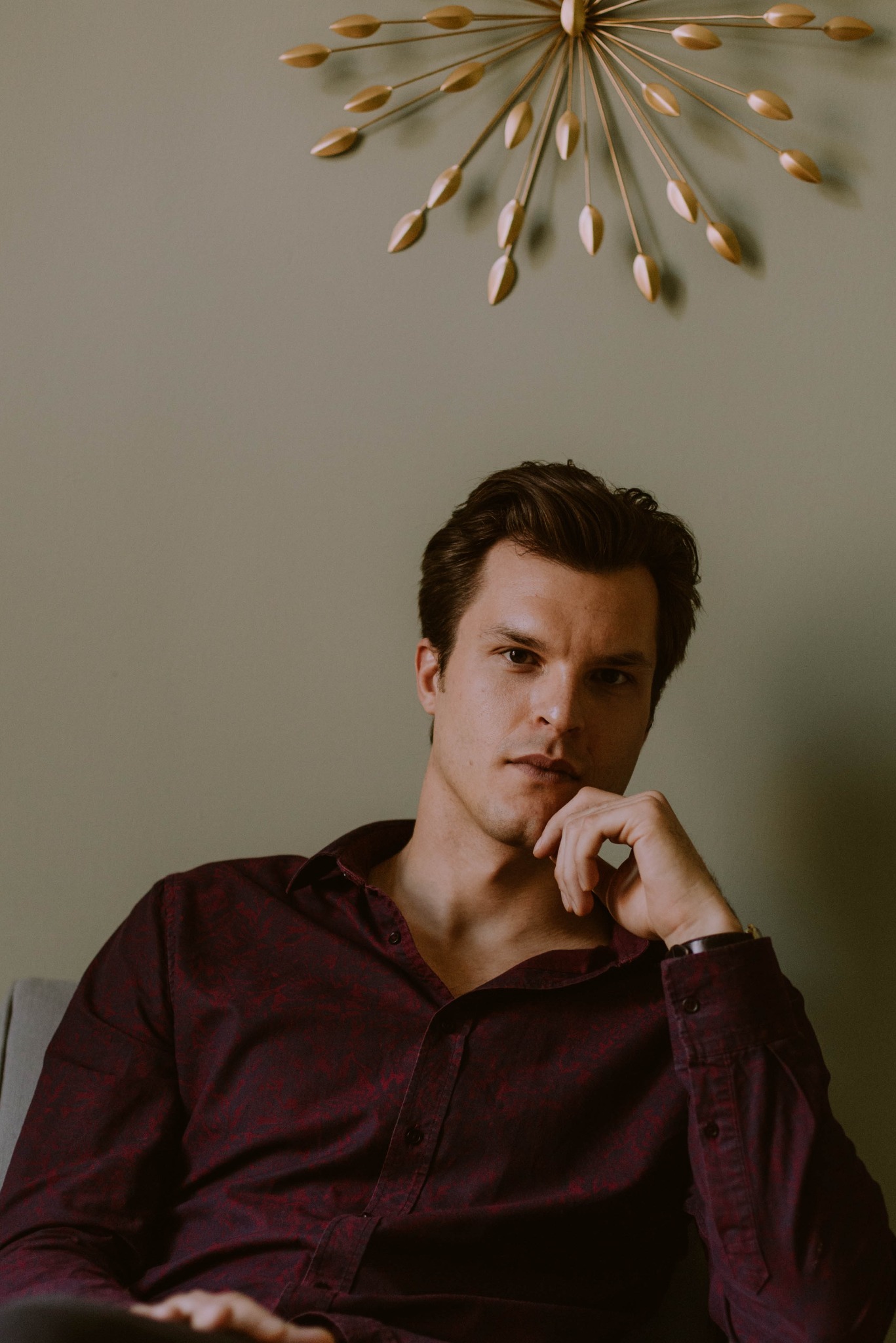 Portret mężczyzny w bordowej koszuli, z ręką na brodzie, pod złotą dekoracją ścienną w kształcie słońca.