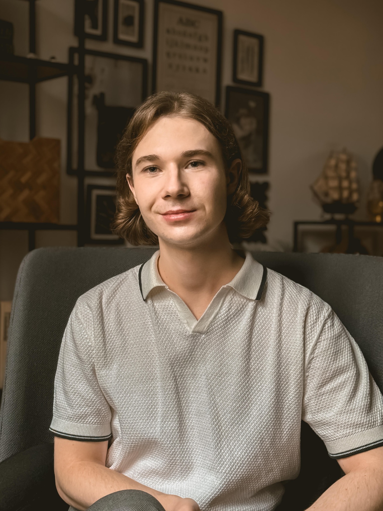 Portret uśmiechniętego młodego mężczyzny z brązowymi włosami siedzącego w szarym fotelu na tle półek z dekoracjami, w tym model żaglowca.