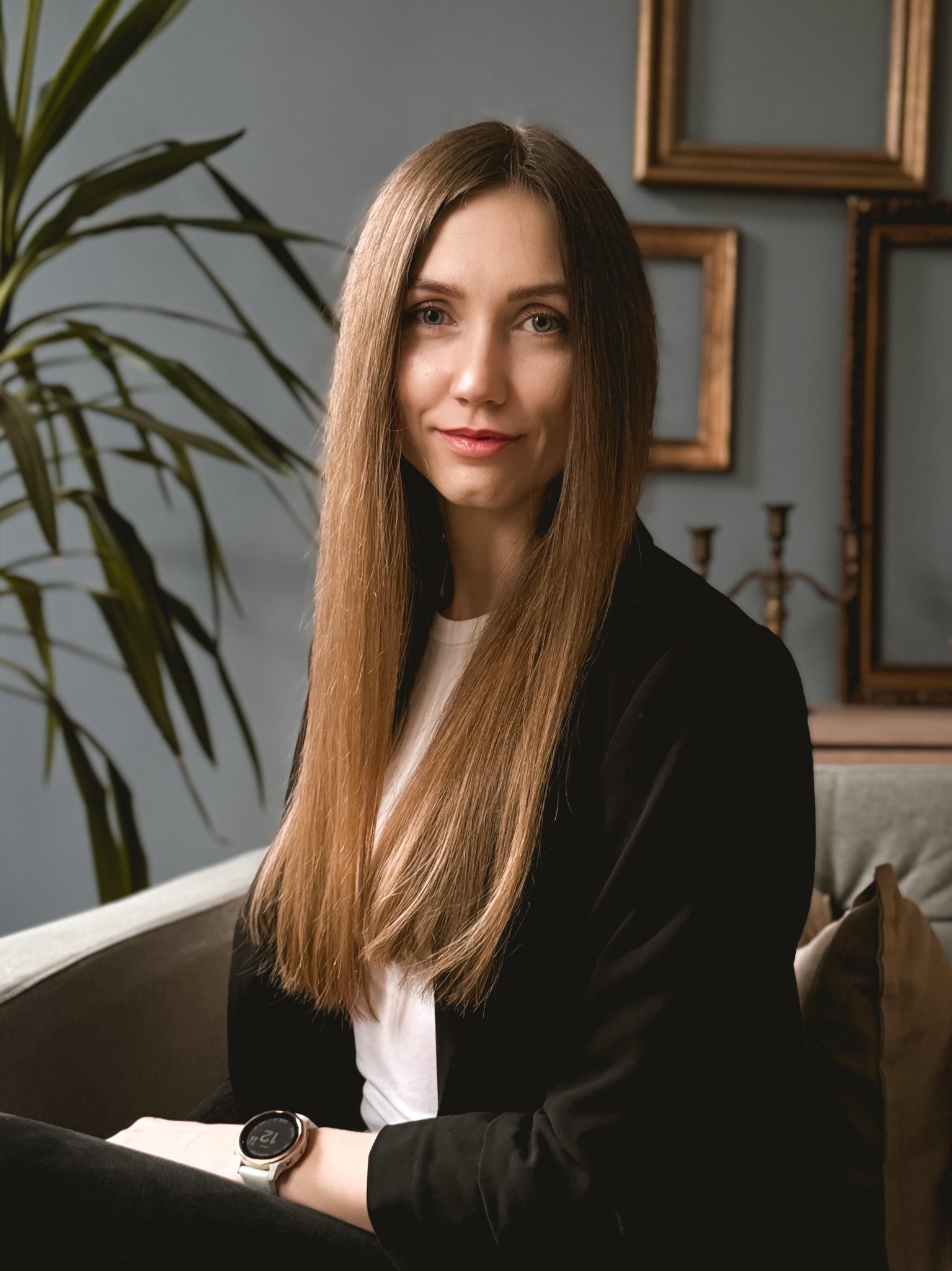 Portret uśmiechniętej psychodietetyczki z Poznania, Moniki Grzelak, w czarnej marynarce, na tle rośliny doniczkowej i dekoracyjnych ramek na ścianie. Widoczny zegarek na ręce.