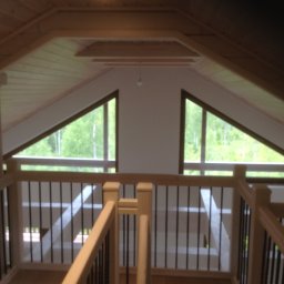 FIRMA STOLARSKO-CIESIELSKA PIOTR KUBIK - Wnętrze domu z drewnianym stropem i balustradą z czarnymi prętami, widok na zieleń przez trójkątne okna.