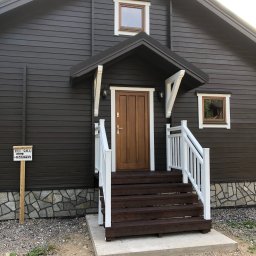 FIRMA STOLARSKO-CIESIELSKA PIOTR KUBIK - Ciemnobrązowy dom z drewnianymi schodami, białymi balustradami i daszkiem nad wejściem, obok tabliczka z napisem 'TILL SALU' i numerem telefonu.