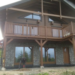 FIRMA STOLARSKO-CIESIELSKA PIOTR KUBIK - Dwukondygnacyjny dom z drewnianą elewacją i kamienną podmurówką, z drewnianym balkonem na piętrze. Robotnik z łopatą przygotowuje grunt przed budynkiem.