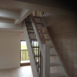 FIRMA STOLARSKO-CIESIELSKA PIOTR KUBIK - Jasne, drewniane schody z czarnymi balustradami w nowym domu, widok z dołu na górę, okno w tle.