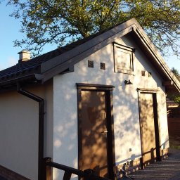 FIRMA STOLARSKO-CIESIELSKA PIOTR KUBIK - Mały budynek gospodarczy z dwiema brązowymi drzwiami, wykończony w jasnej kolorystyce, z ciemnym dachem i widocznymi elementami orynnowania, otoczony zielenią drzew i kostką brukową.