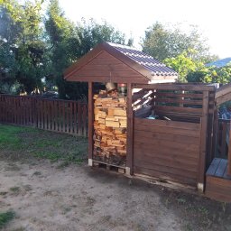 FIRMA STOLARSKO-CIESIELSKA PIOTR KUBIK - Drewniana konstrukcja z zadaszeniem wypełniona pociętym drewnem na opał, obok widoczny drewniany płot w ogrodzie.