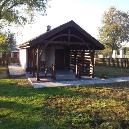 FIRMA STOLARSKO-CIESIELSKA PIOTR KUBIK - Drewniana konstrukcja z dachem krytym dachówką, pełniąca funkcję zadaszenia tarasu lub altany, z elementami ażurowymi i kolumnami, połączona z budynkiem mieszkalnym i otoczona zielonym trawnikiem...