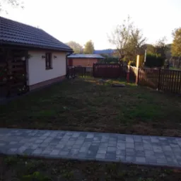 Widok na posesję z drewnianym ogrodzeniem w kolorze ciemnego brązu, trawnik w trakcie renowacji, dom z dekoracyjną, drewnianą balustradą przy tarasie i wybrukowanym chodnikiem z szarych płyt.