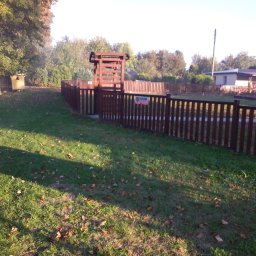 FIRMA STOLARSKO-CIESIELSKA PIOTR KUBIK - Ciemnobrązowe ogrodzenie z drewnianych sztachet otacza trawnik z opadłymi liśćmi, widoczny fragment drewnianej konstrukcji nad bramą i tabliczka z logo firmy ochroniarskiej.