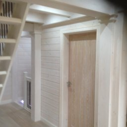 FIRMA STOLARSKO-CIESIELSKA PIOTR KUBIK - Wnętrze domu z drewnianą boazerią pomalowaną na biało, widoczne schody, drewniane drzwi wewnętrzne i fragment antresoli z drewnianymi belkami stropowymi.