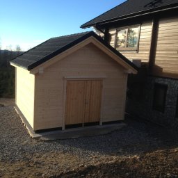 FIRMA STOLARSKO-CIESIELSKA PIOTR KUBIK - Mały, surowy, drewniany budynek gospodarczy z dwuspadowym dachem, stojący na żwirowym podłożu obok większego domu z elementami drewnianymi i kamiennymi.