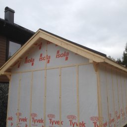 FIRMA STOLARSKO-CIESIELSKA PIOTR KUBIK - Konstrukcja szkieletowa drewnianej dobudówki z widoczną membraną izolacyjną z nadrukiem logo producentów, na tle domu z brązową elewacją i szarym niebem.