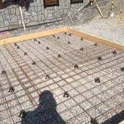 FIRMA STOLARSKO-CIESIELSKA PIOTR KUBIK - Przygotowanie podłoża pod konstrukcję: drewniana rama, siatka zbrojeniowa z dystansami na żwirowym podłożu, cień osoby w kadrze.