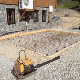 FIRMA STOLARSKO-CIESIELSKA PIOTR KUBIK - Przygotowanie podłoża pod przyszły taras: siatka zbrojeniowa, drewniany szalunek i ubijarka wibracyjna na żwirowym podłożu przed budynkiem z drewnianą elewacją i kamienną podmurówką.