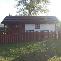 FIRMA STOLARSKO-CIESIELSKA PIOTR KUBIK - Ogród z domkiem letniskowym, drewnianym ogrodzeniem i zadaszeniem tarasu, widok w słoneczny dzień.