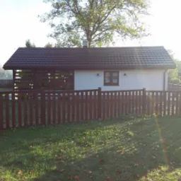 Ogród z domkiem letniskowym, drewnianym ogrodzeniem i zadaszeniem tarasu, widok w słoneczny dzień.