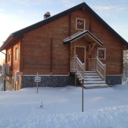 FIRMA STOLARSKO-CIESIELSKA PIOTR KUBIK - Drewniany dom z drewnianymi schodami wejściowymi, pokryty śniegiem, z tabliczką informacyjną 'TILL SALU'.