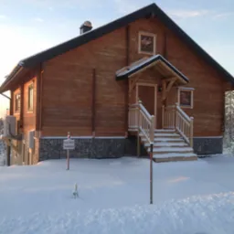 Drewniany dom z drewnianymi schodami wejściowymi, pokryty śniegiem, z tabliczką informacyjną 'TILL SALU'.