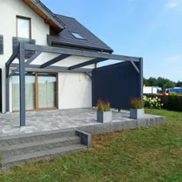 Nowoczesny taras z szarej kostki brukowej, zadaszony konstrukcją z ciemnego drewna przylegającą do budynku z oknem i szklaną balustradą, ozdobiony donicami z trawami ozdobnymi.