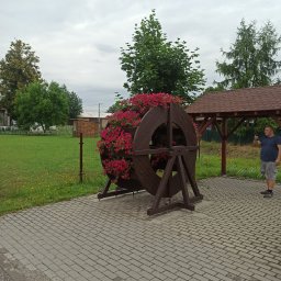 FIRMA STOLARSKO-CIESIELSKA PIOTR KUBIK - Ozdobna, drewniana konstrukcja w kształcie koła z wbudowanymi donicami wypełnionymi czerwonymi kwiatami, umieszczona na zewnątrz na brukowanym terenie, z widocznym mężczyzną robiącym zdjęcie...