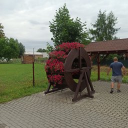 FIRMA STOLARSKO-CIESIELSKA PIOTR KUBIK - Ozdobna, drewniana konstrukcja w kształcie koła, wypełniona czerwonymi kwiatami, ustawiona na brukowanym chodniku w przestrzeni publicznej. W tle widoczny mężczyzna oddalający się od obiektu...