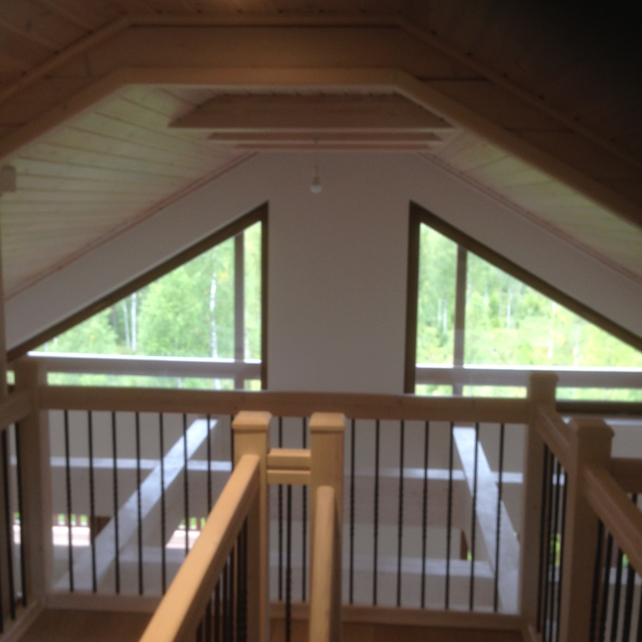 Wnętrze domu z drewnianym stropem i balustradą z czarnymi prętami, widok na zieleń przez trójkątne okna.