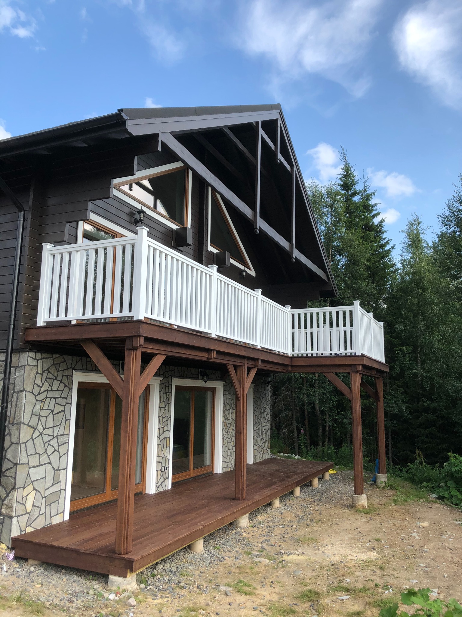 Dwupoziomowy taras przy domu z drewnianą podłogą i białymi balustradami, wsparty na drewnianych kolumnach, z widokiem na las.