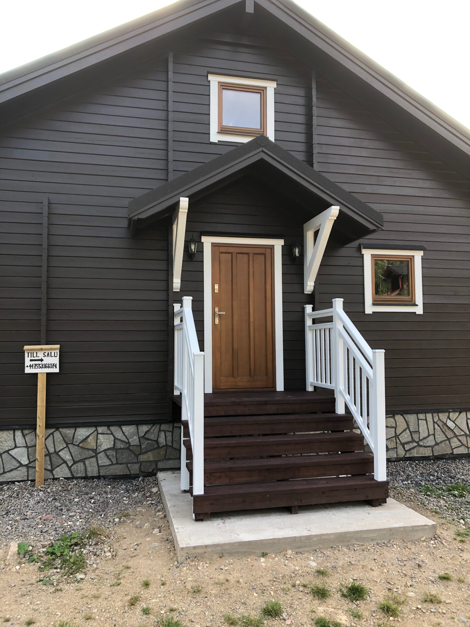 Ciemnobrązowy dom z drewnianymi schodami, białymi balustradami i daszkiem nad wejściem, obok tabliczka z napisem 'TILL SALU' i numerem telefonu.