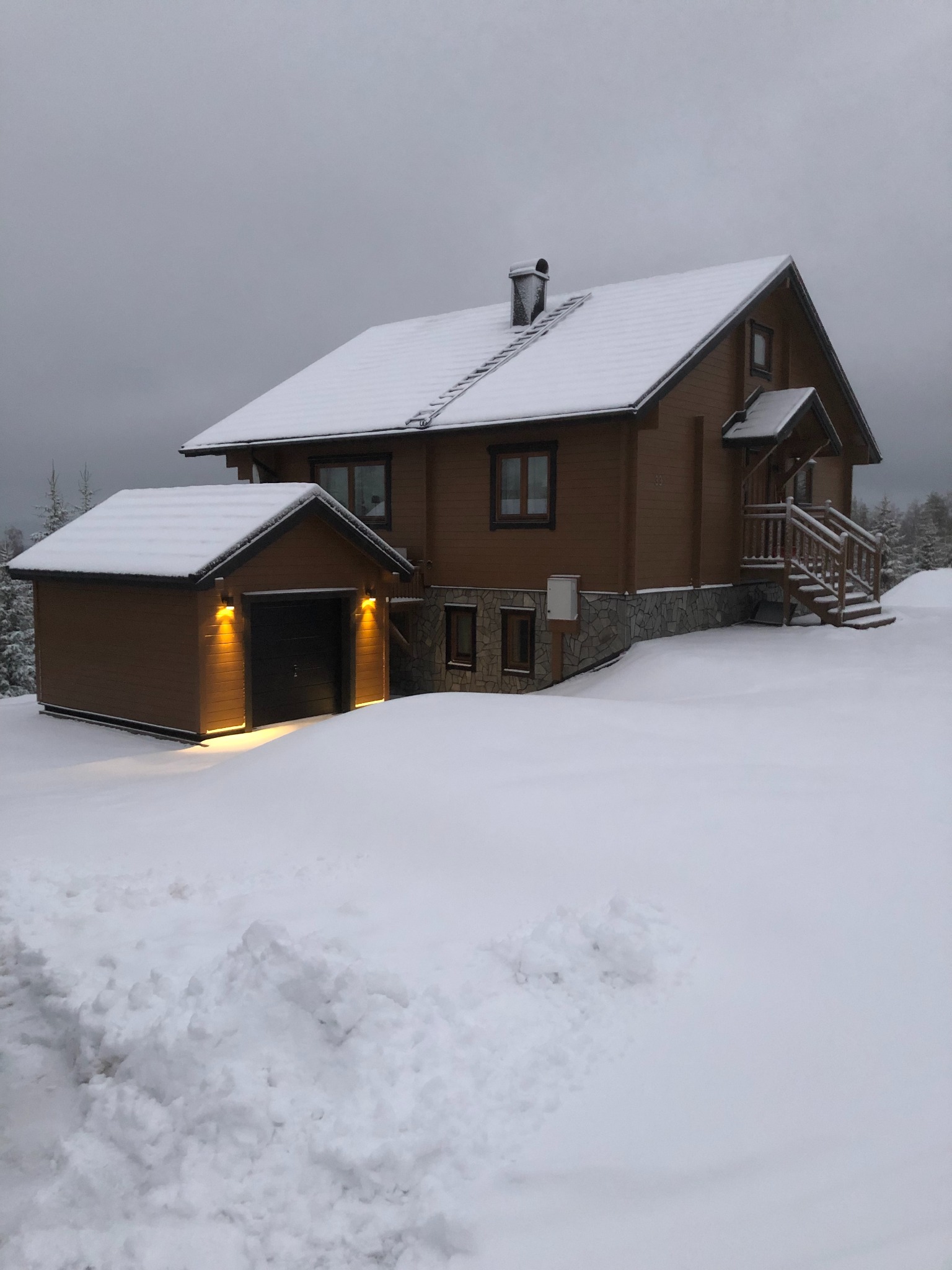 Zimowy krajobraz z drewnianym domem i garażem, oba pokryte śniegiem, schody wejściowe z drewnianą balustradą również pod śniegiem, pochmurne niebo w tle.