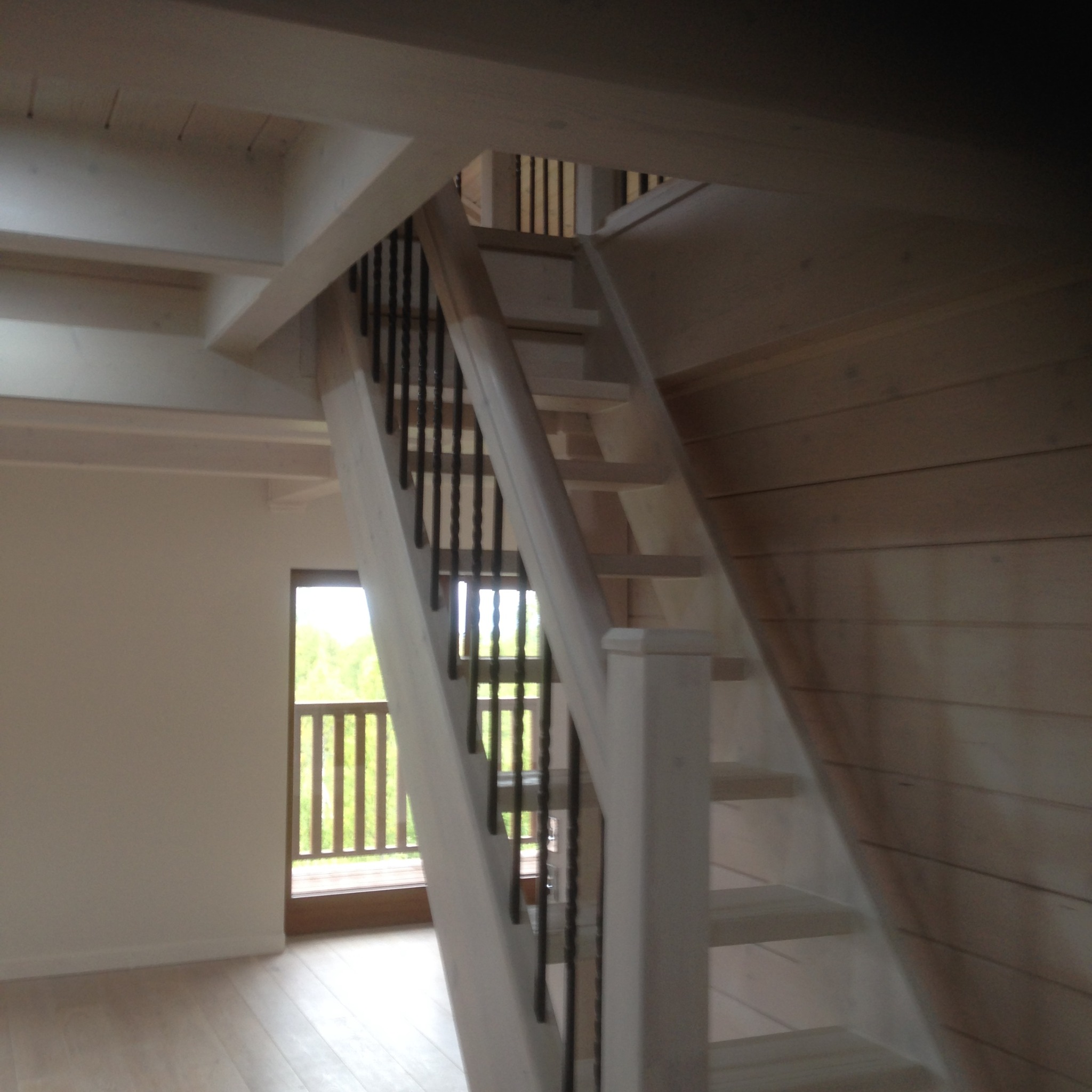 Jasne, drewniane schody z czarnymi balustradami w nowym domu, widok z dołu na górę, okno w tle.