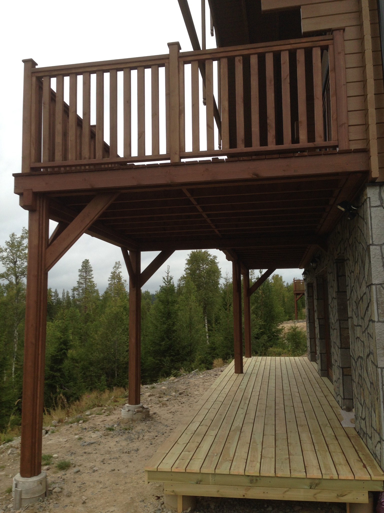 Wielopoziomowy, drewniany taras z balustradą, wsparty na solidnych filarach, z widokiem na las w pochmurny dzień.