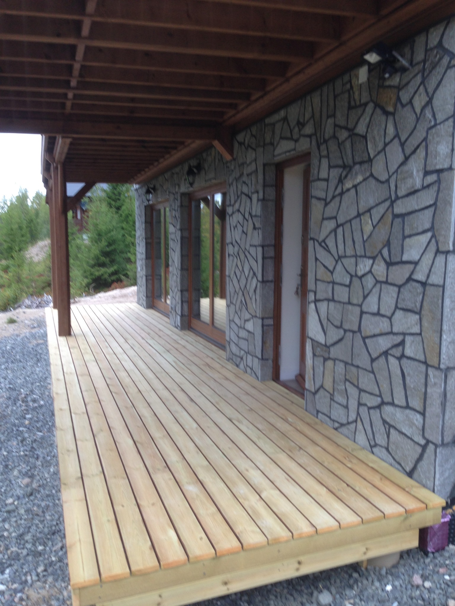 Zadaszony taras z jasnego drewna, przylegający do kamiennego budynku z trzema oknami i drzwiami, otoczony żwirowym podłożem i zielenią.