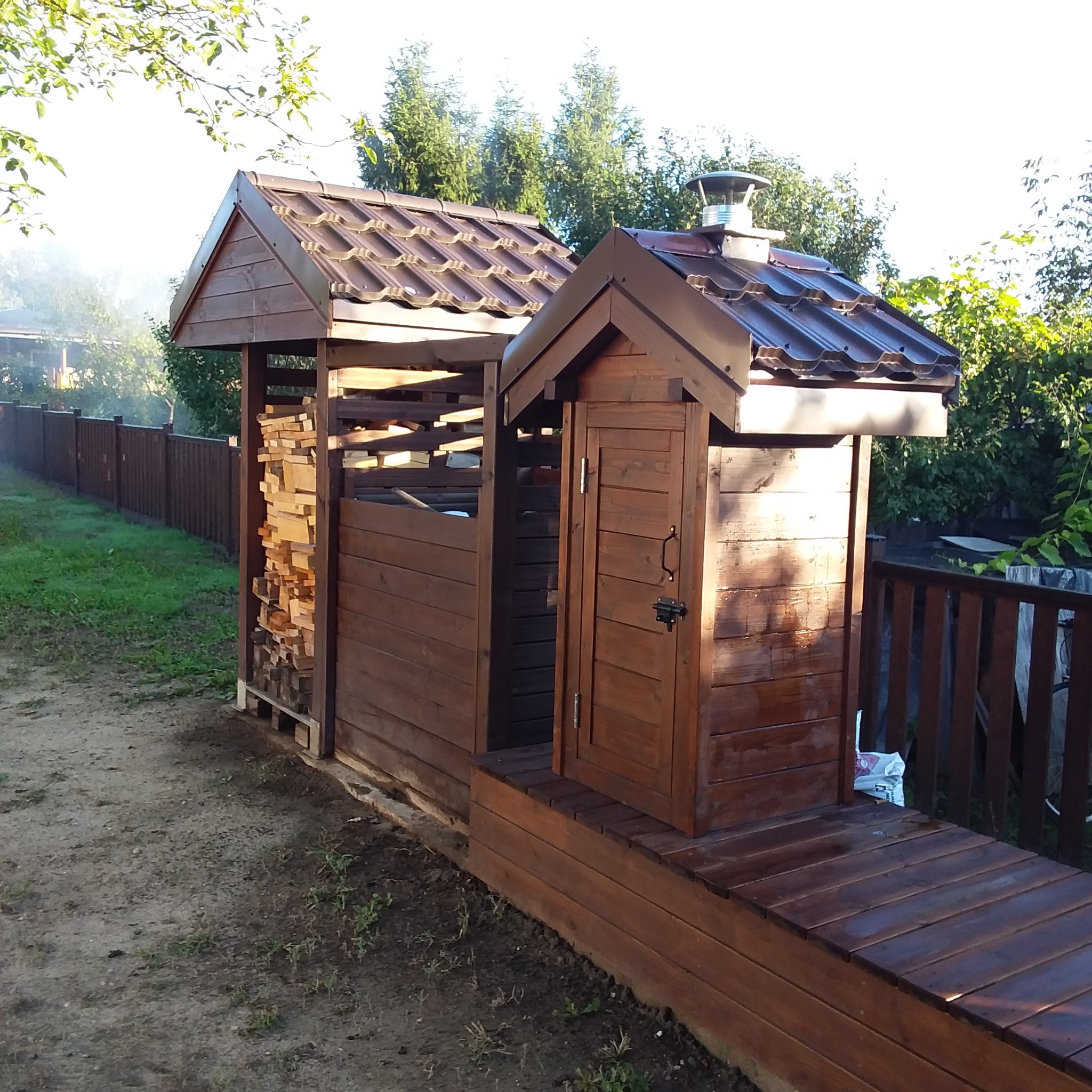 Drewniana drewutnia z zamykaną szafką, brązowy dach z blachodachówki, widok z zewnątrz, obok drewniany płot.