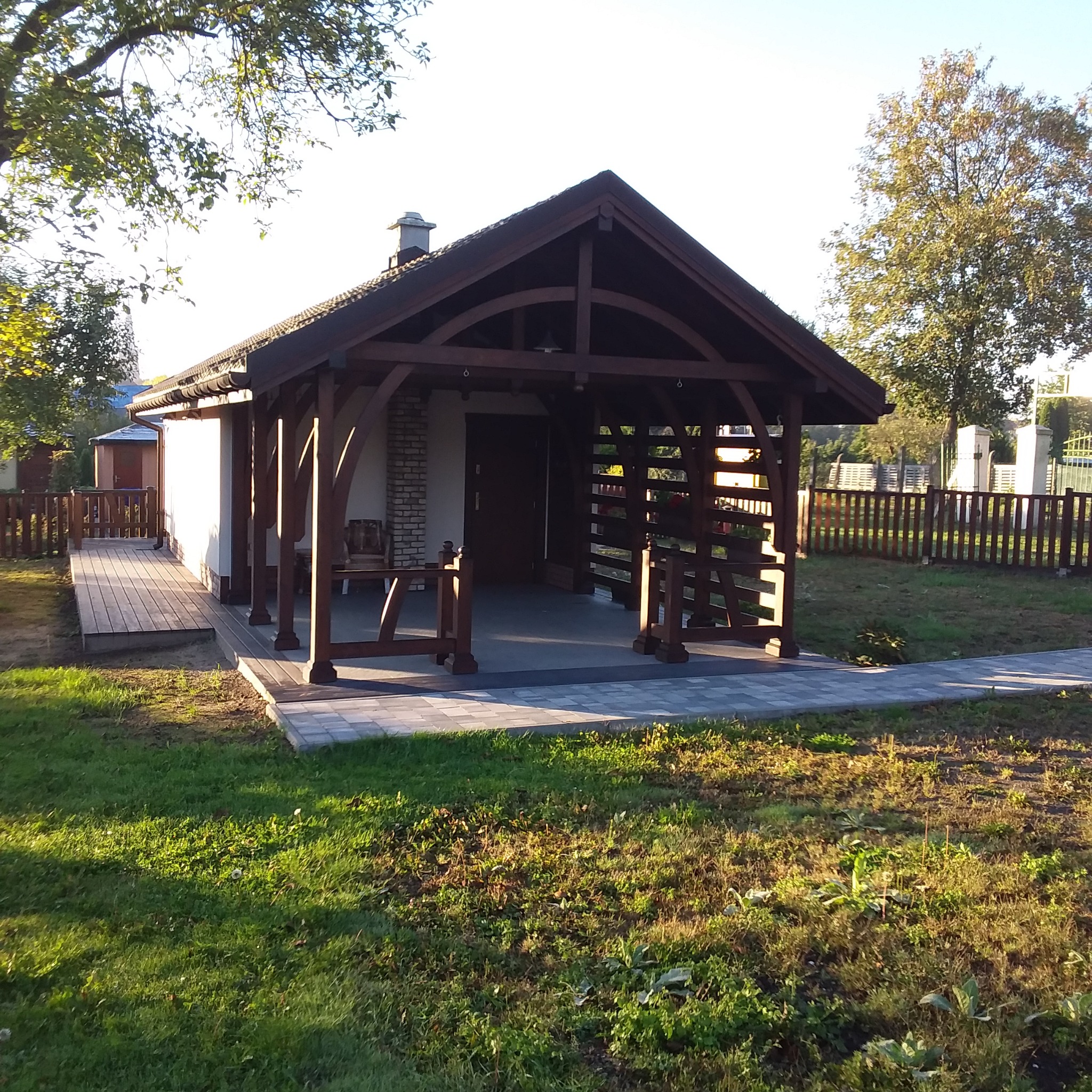 Drewniana konstrukcja z dachem krytym dachówką, pełniąca funkcję zadaszenia tarasu lub altany, z elementami ażurowymi i kolumnami, połączona z budynkiem mieszkalnym i otoczona zielonym trawnikiem...