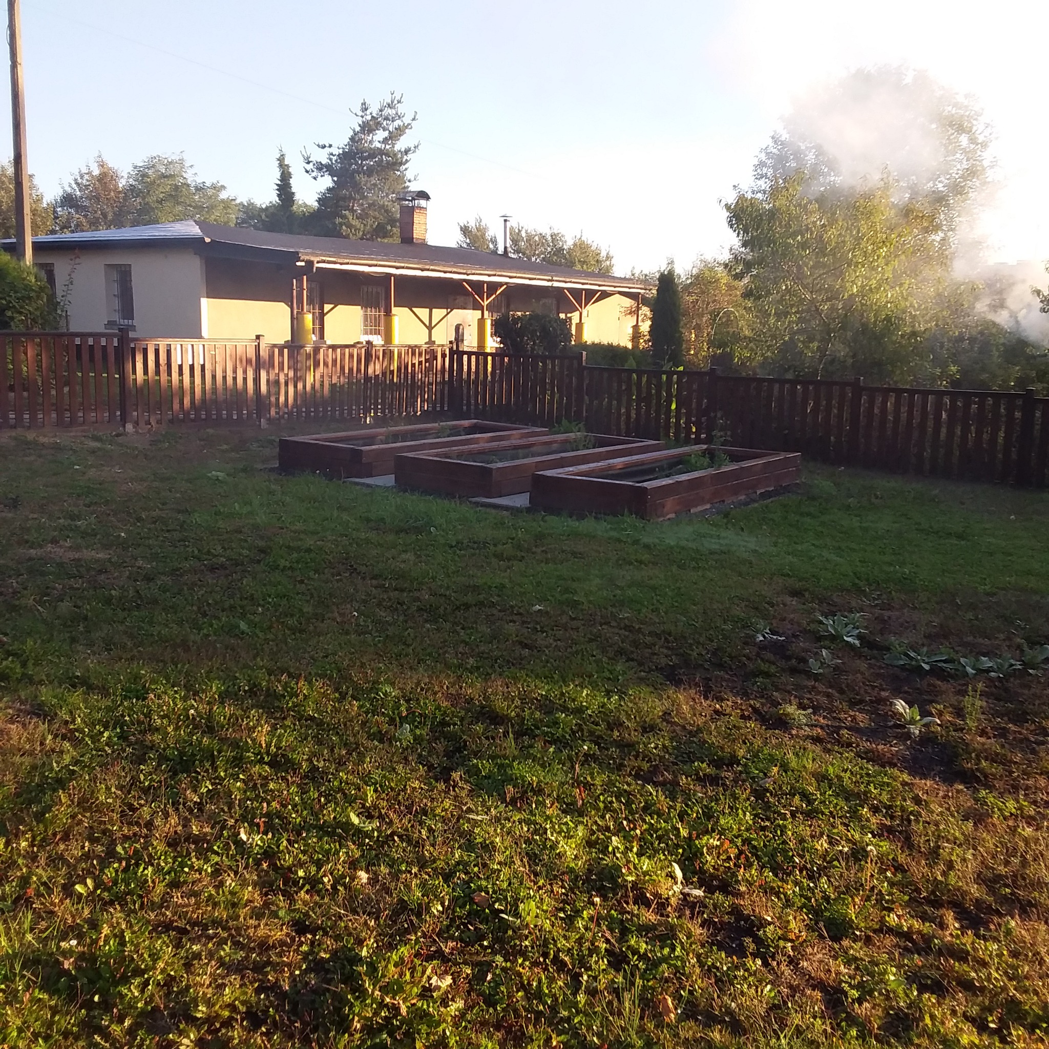 Widok na ogród z trzema drewnianymi podwyższonymi grządkami, otoczonymi drewnianym ogrodzeniem, z domem z zadaszonym tarasem w tle i dymem unoszącym się zza drzew