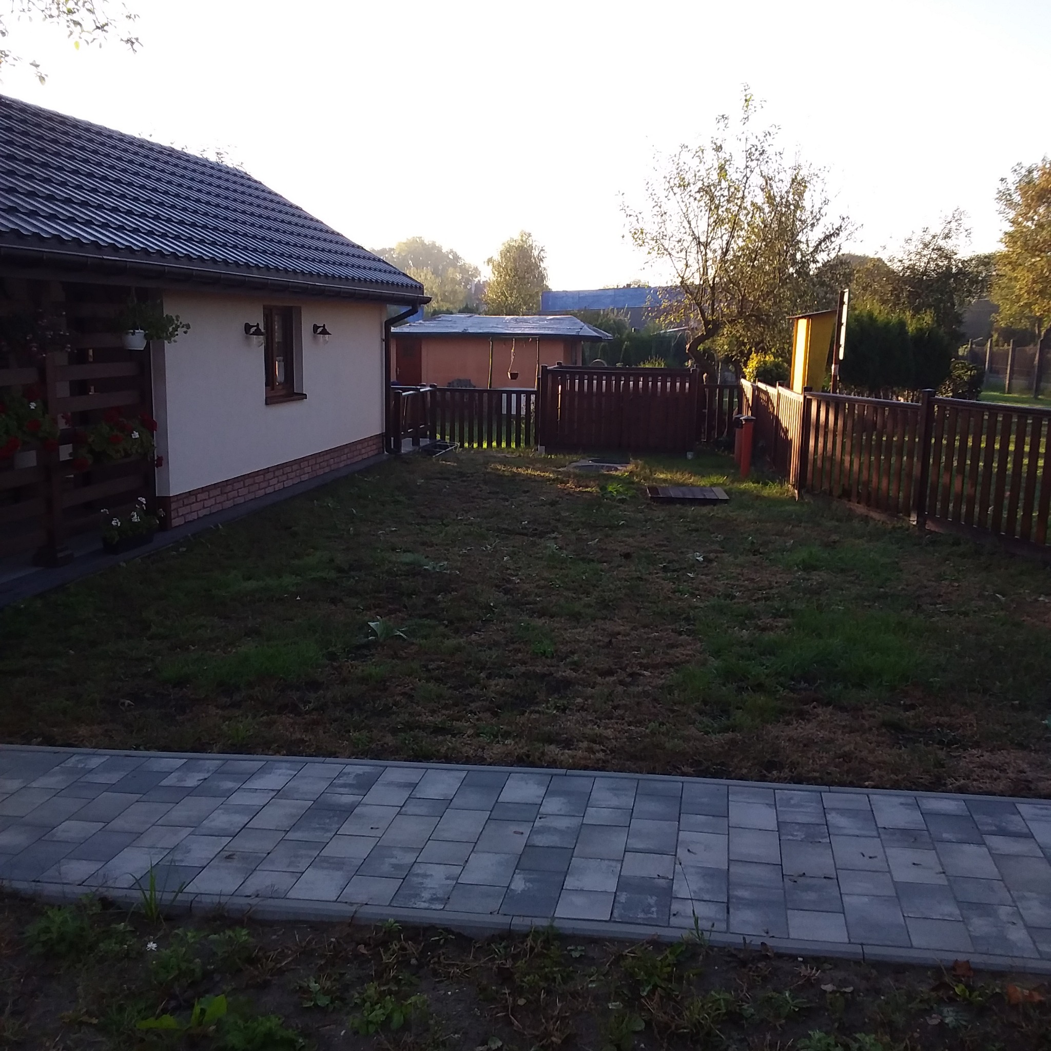 Widok na posesję z drewnianym ogrodzeniem w kolorze ciemnego brązu, trawnik w trakcie renowacji, dom z dekoracyjną, drewnianą balustradą przy tarasie i wybrukowanym chodnikiem z szarych płyt.