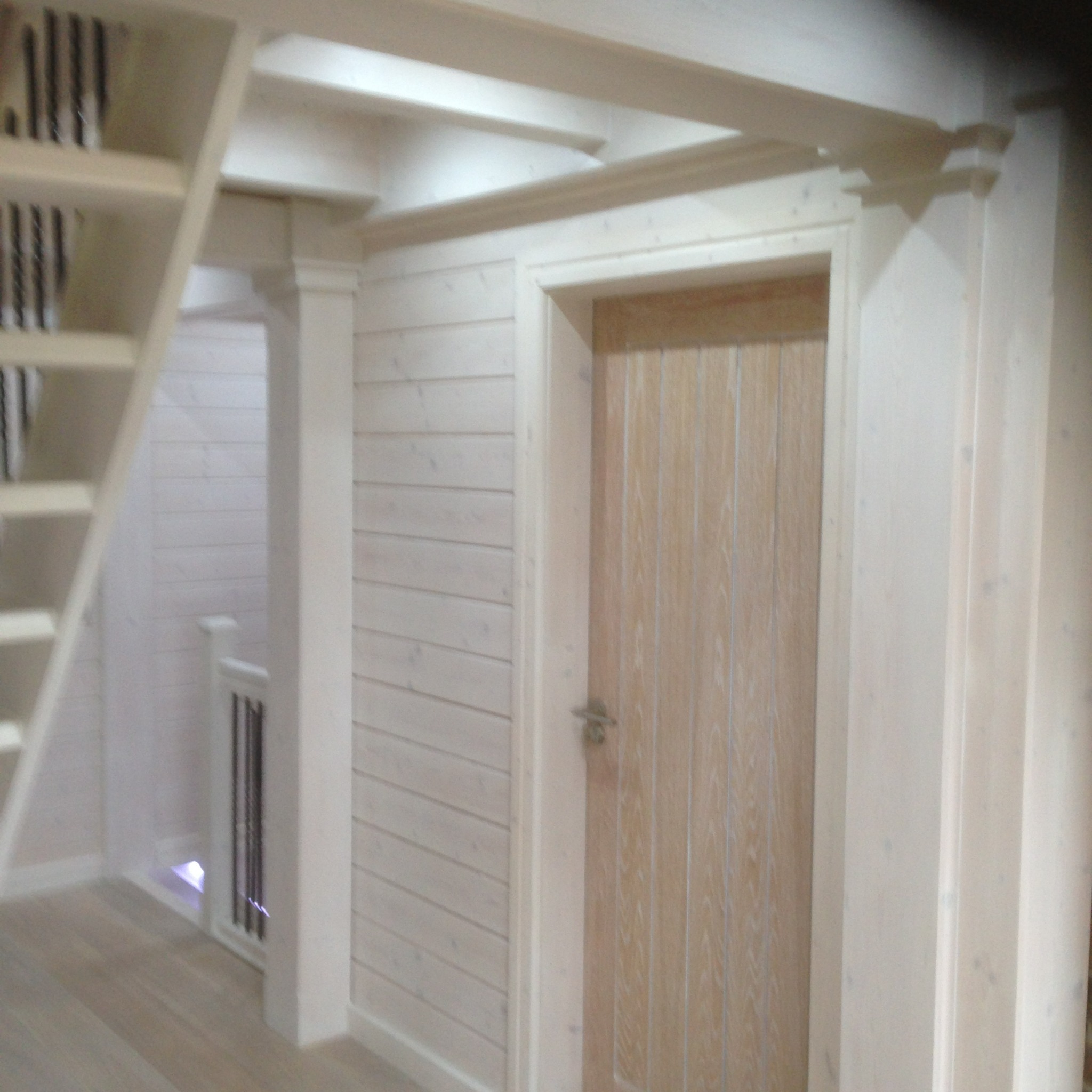 Wnętrze domu z drewnianą boazerią pomalowaną na biało, widoczne schody, drewniane drzwi wewnętrzne i fragment antresoli z drewnianymi belkami stropowymi.