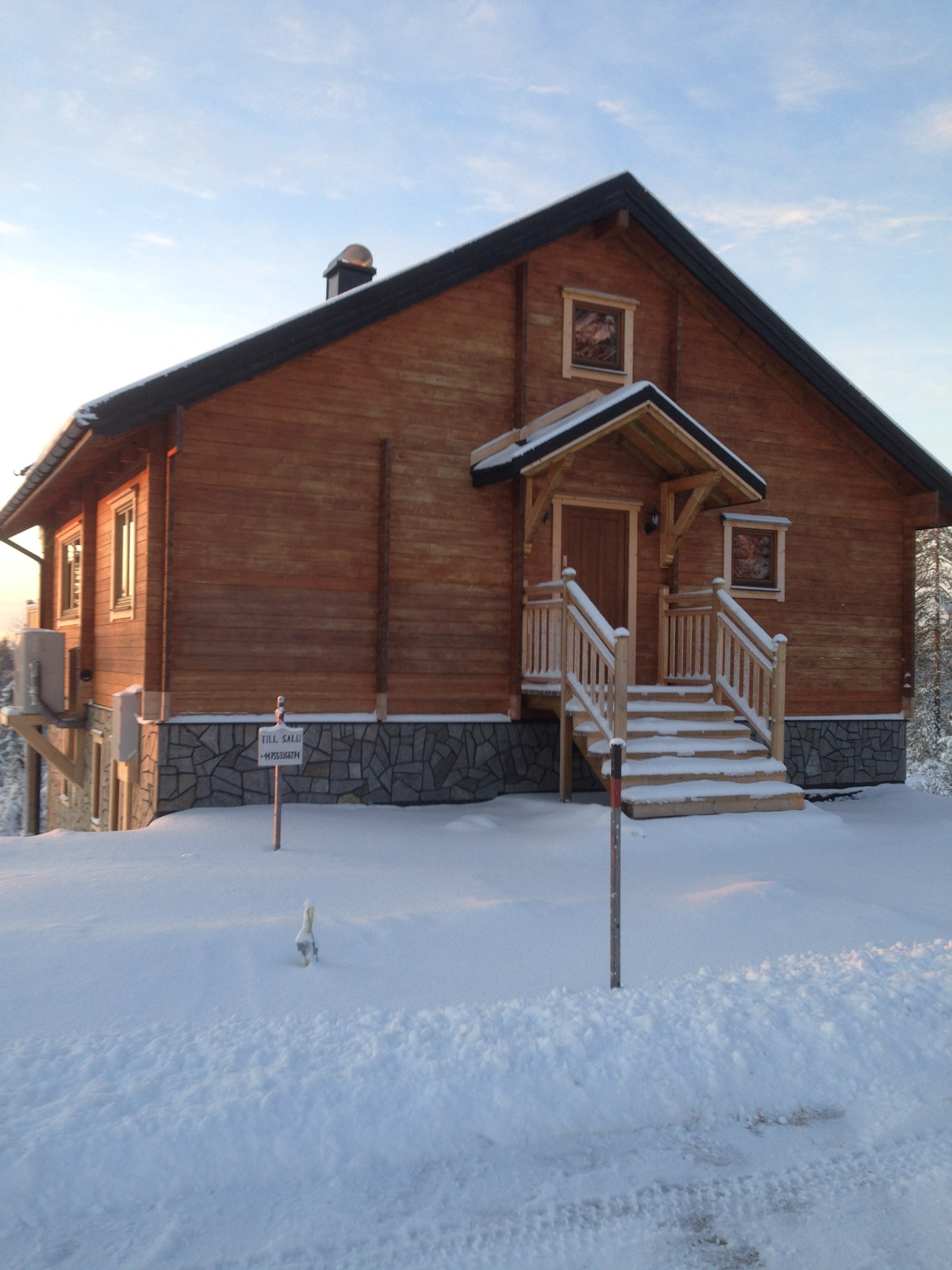 Drewniany dom z drewnianymi schodami wejściowymi, pokryty śniegiem, z tabliczką informacyjną 'TILL SALU'.