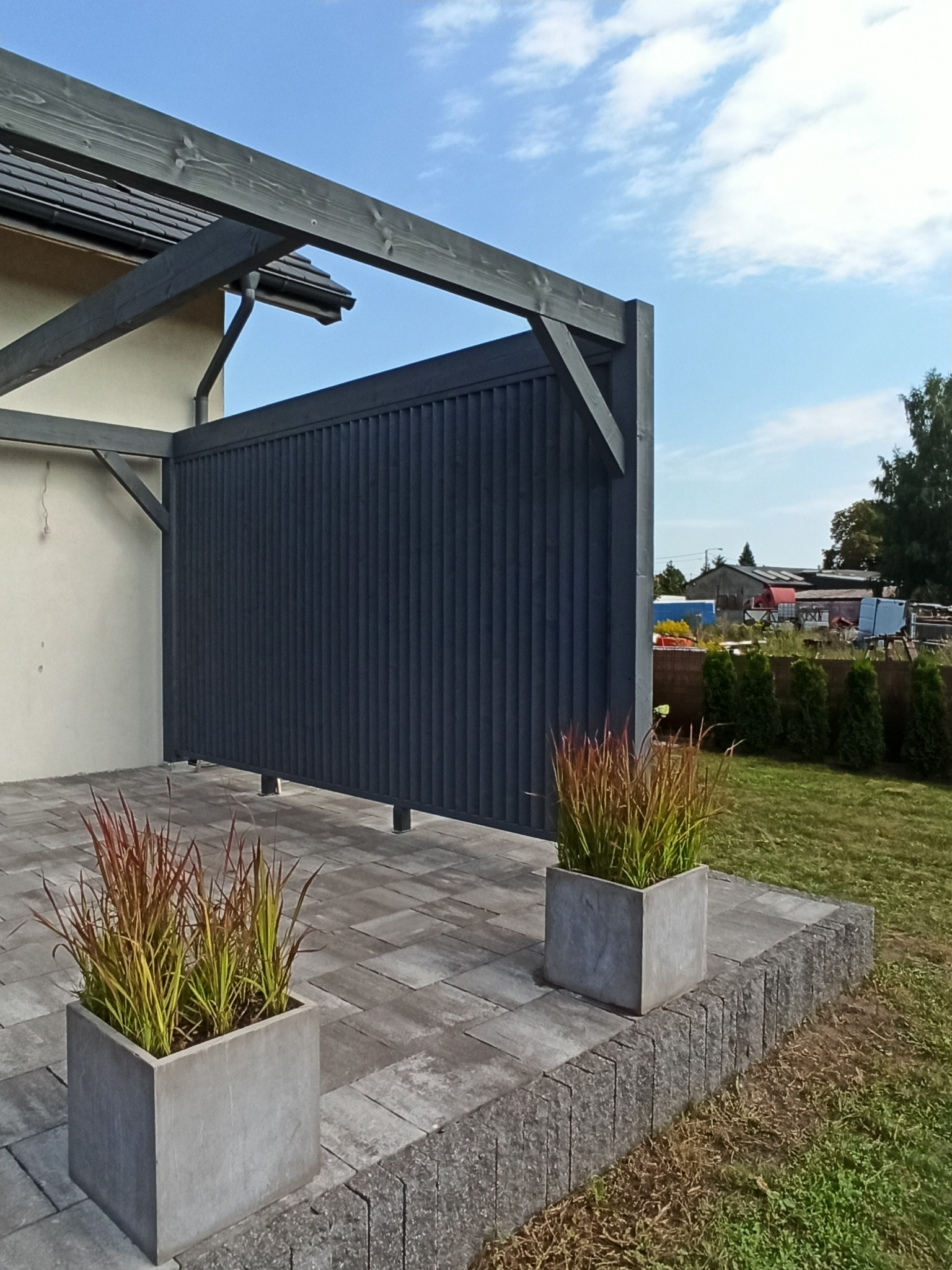Nowoczesna konstrukcja z ciemnego drewna, prawdopodobnie osłona tarasu lub element pergoli, z widocznymi donicami z ozdobnymi trawami na brukowanym podłożu.