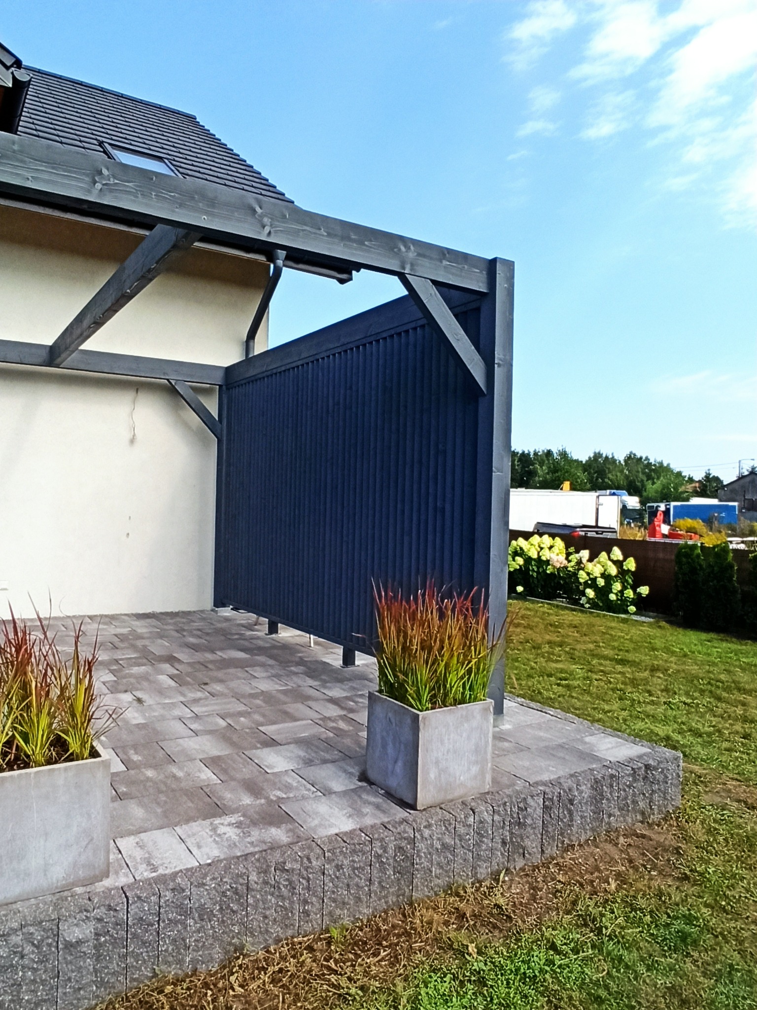 Zadaszony taras z ciemnego drewna, wykończony kostką brukową i ozdobiony donicami z trawami ozdobnymi, na tle domu z szarą dachówką i błękitnego nieba.