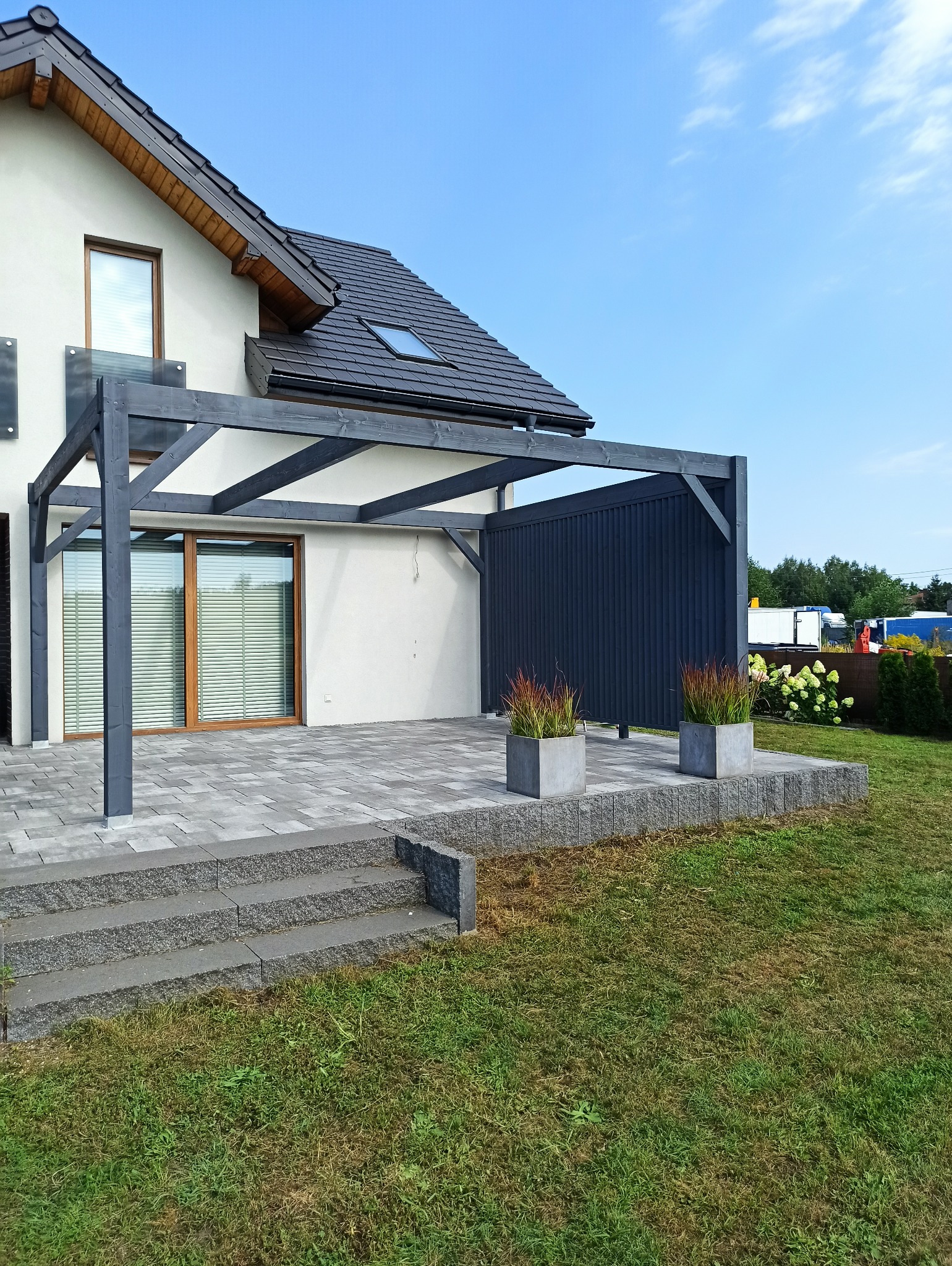 Nowoczesny taras z szarej kostki brukowej, zadaszony konstrukcją z ciemnego drewna przylegającą do budynku z oknem i szklaną balustradą, ozdobiony donicami z trawami ozdobnymi.