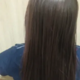 Widok z tyłu na długie, proste, brązowe włosy po zabiegu pielęgnacyjnym, z delikatnym połyskiem i minimalnym rozdwajaniem końcówek, na rozmytym tle ściany.